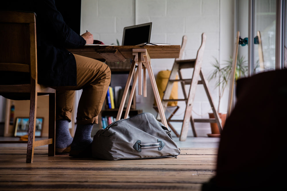 Person sitting at a desk working on a laptop with a Millican Miles the Duffle Bag 28L on the floor beside them