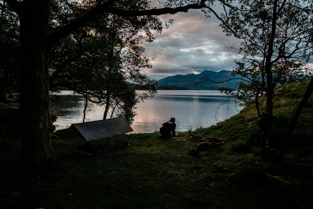 Millican | A person sits by a campfire at a lakeside campsite during dusk, with a tarp shelter among trees on the left and distant mountains under a twilight sky.
