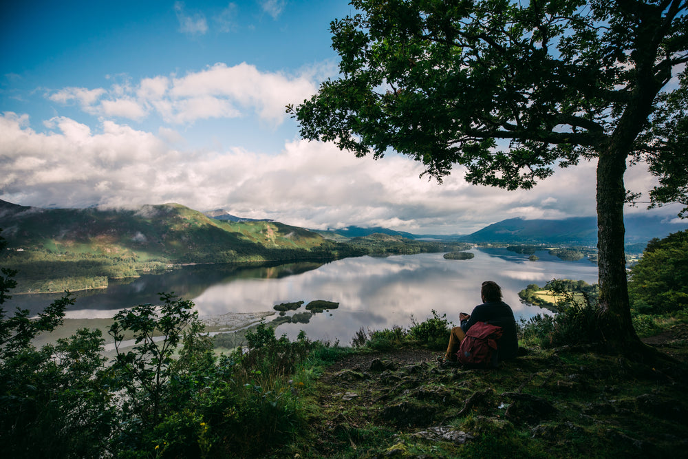 A person sits under a tree on a hill overlooking a calm lake with small islands and surrounded by mountains, with a Millican Fraser 32L backpack beside them