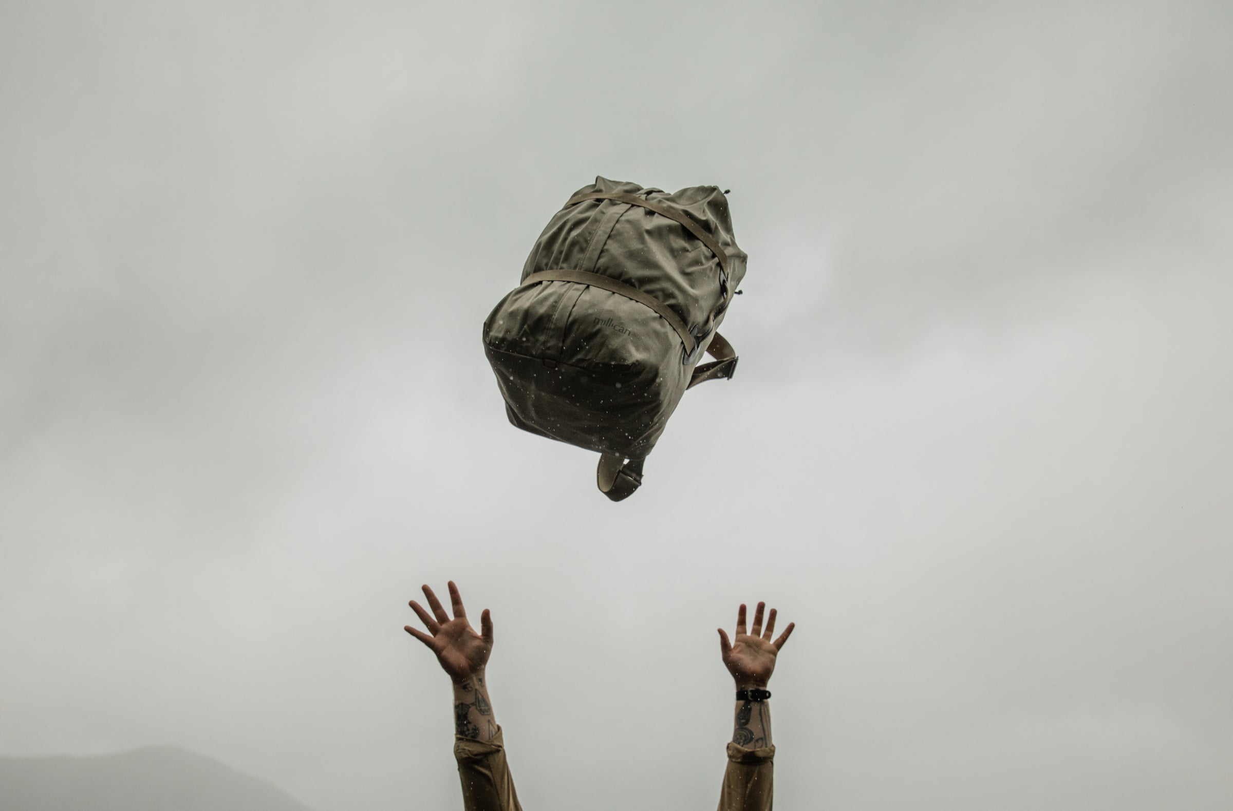 A Millican Miles 40L duffle bag is captured mid-air against a grey sky, with a person's hands reaching up from the bottom of the frame