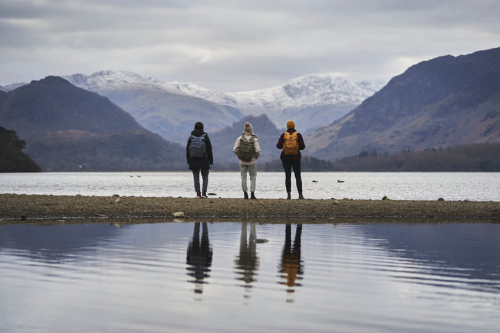 Three people stand by a lake, facing snow-capped mountains, each wearing a Millican Smith Roll backpack