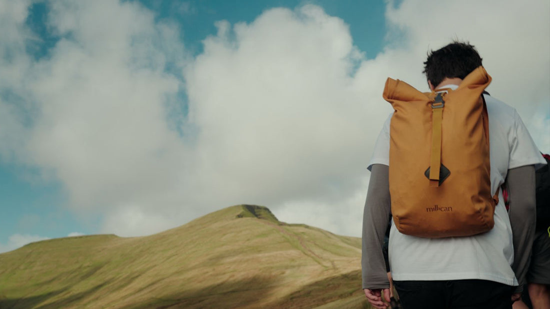 Person hiking with a yellow Millican bag on their back, walking towards a hill under a bright blue sky