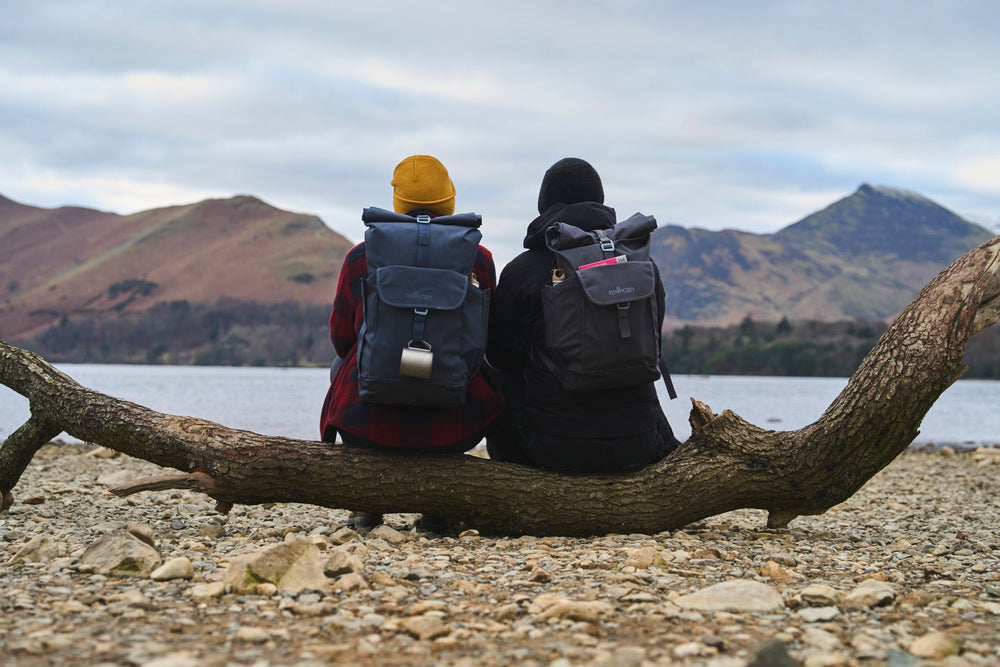 Two people sitting by a lake with Millican backpacks, looking out toward the mountains