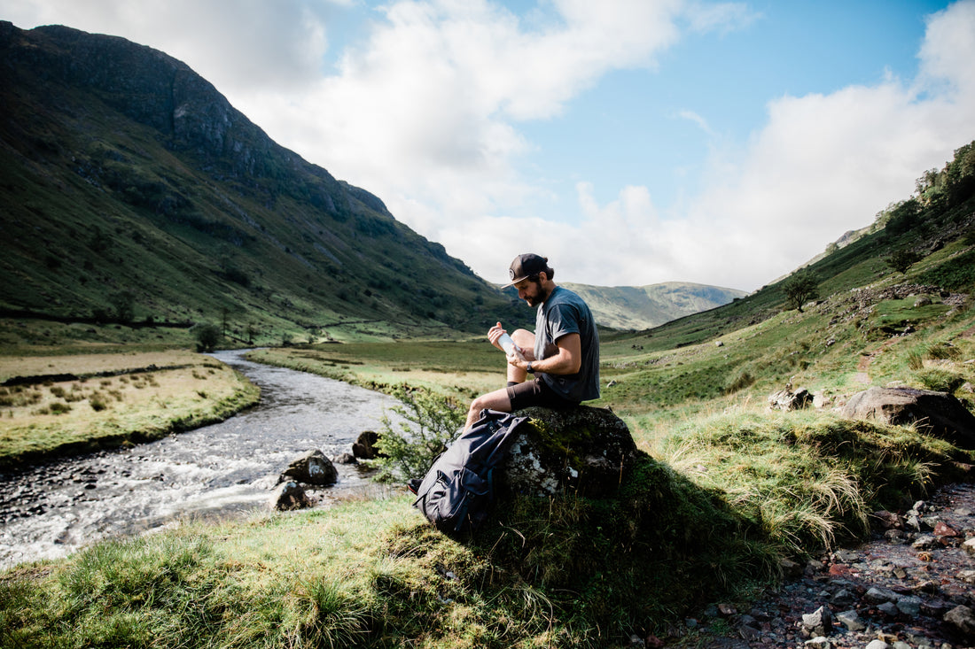 A person wearing a cap sits on a rock near a flowing river in a lush green valley, examining a water bottle. A Millican backpack rests on the ground beside them.