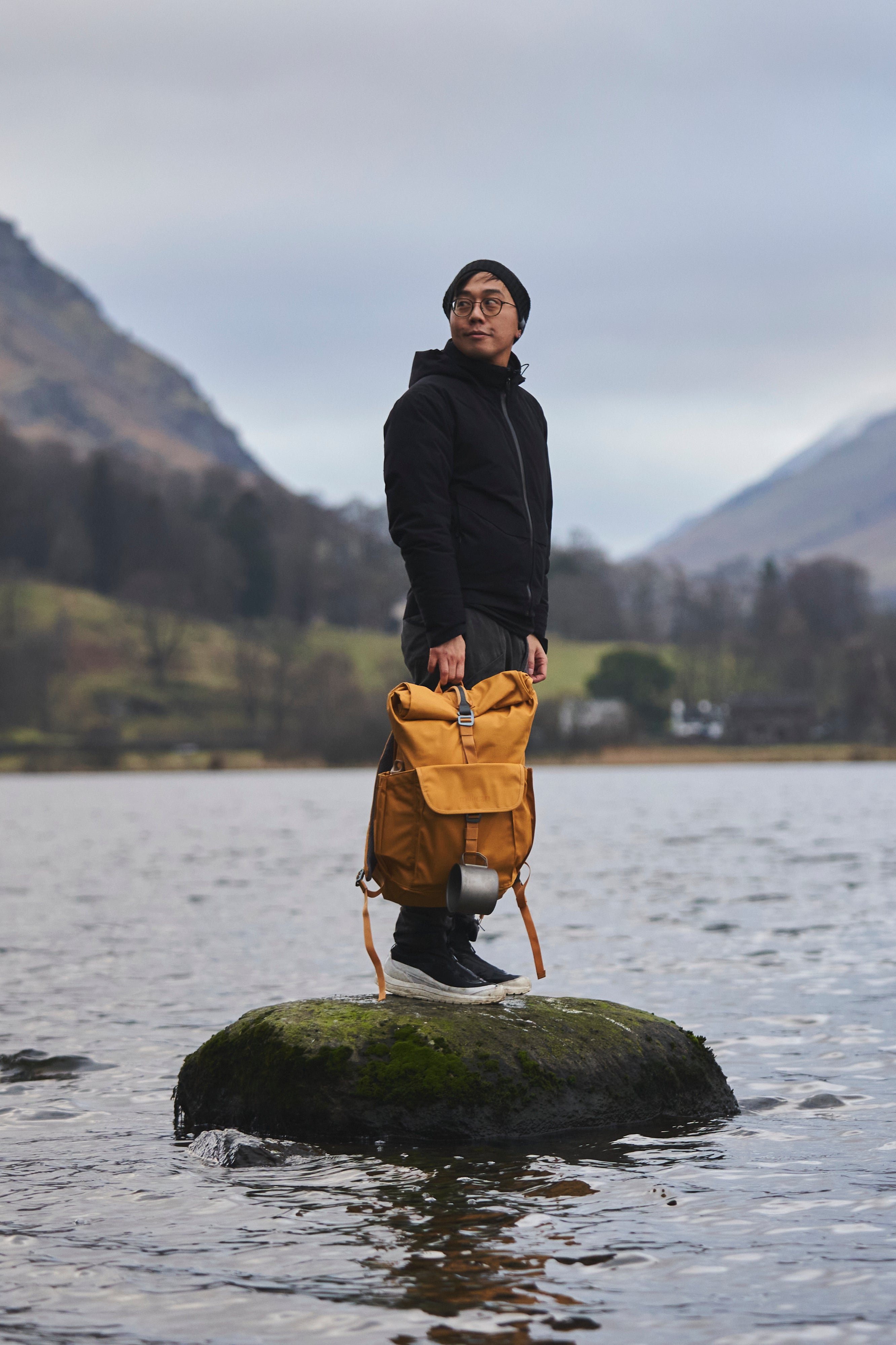 A person standing on a mossy rock in a lake, holding a Gorse Smith the Roll Pack