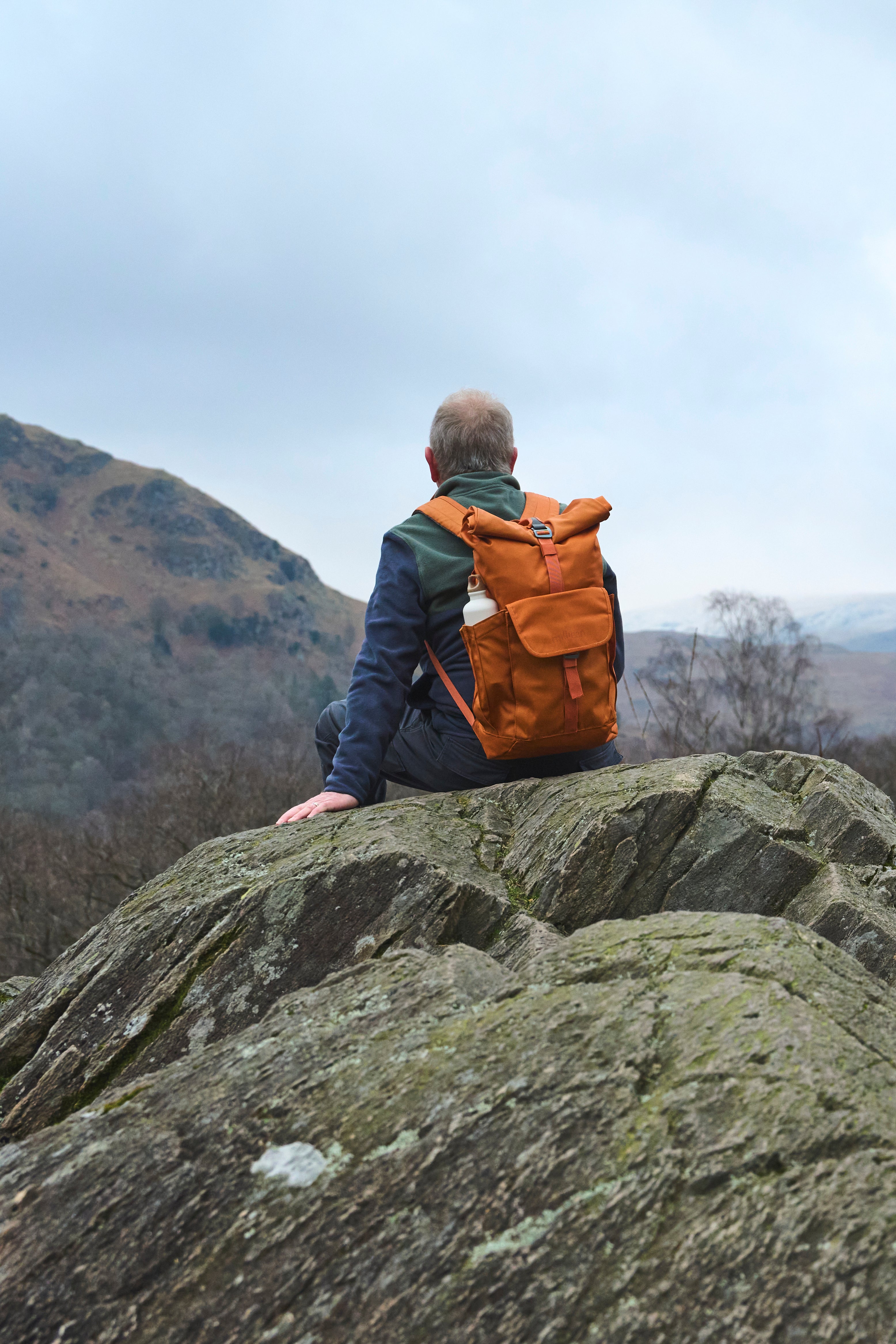 A person sitting on a rocky hill, facing away, wearing an Orange Smith the Roll Pack