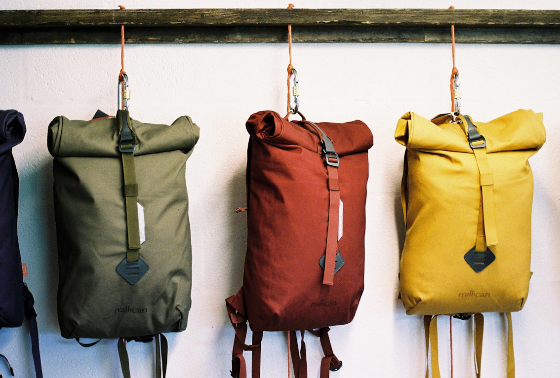 Millican roll-top backpacks hanging side by side on a wooden rack